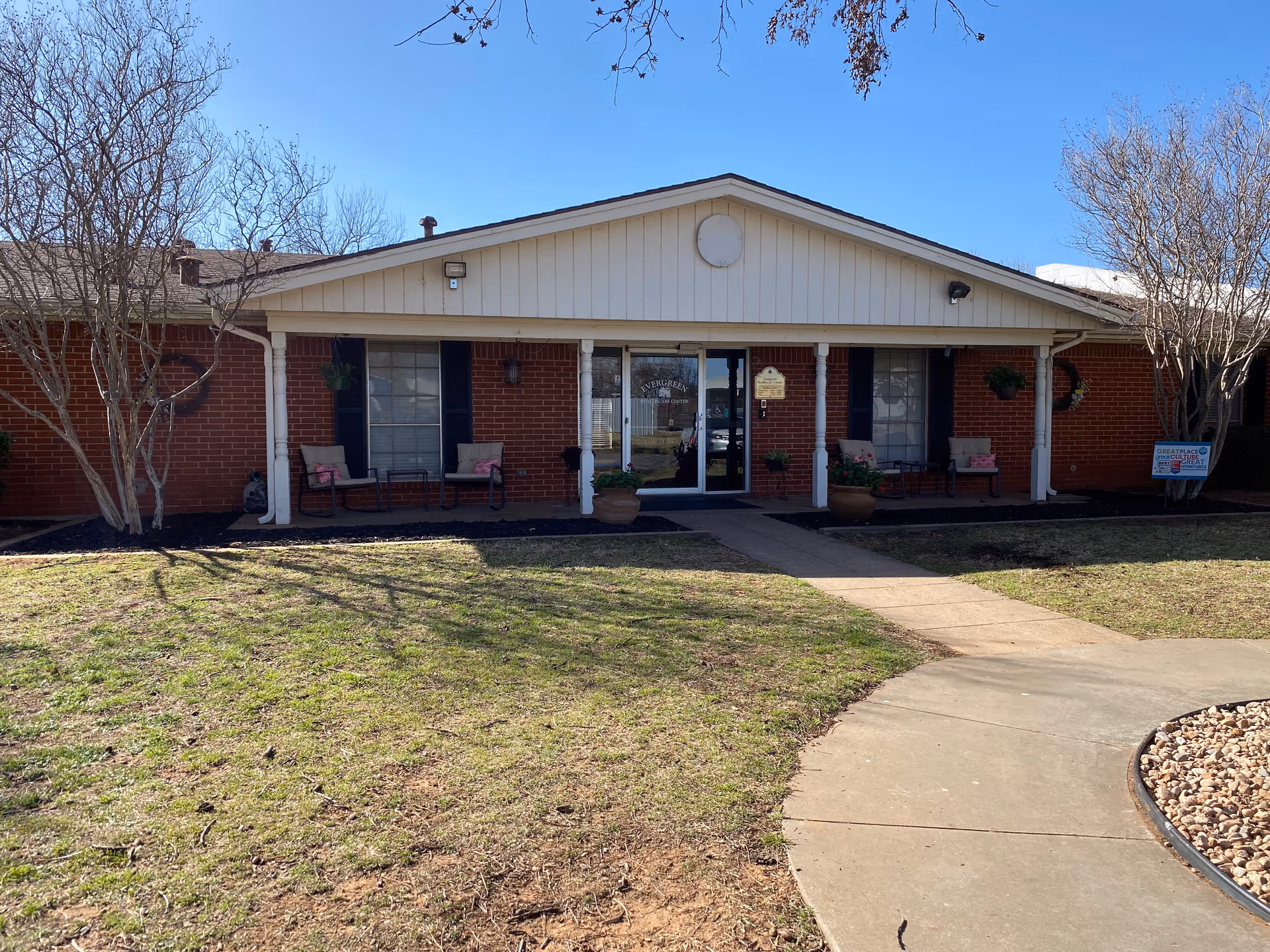 Front entrance of a single-story brick senior care facility with a covered porch, seating and a walkway across a grassy lawn.