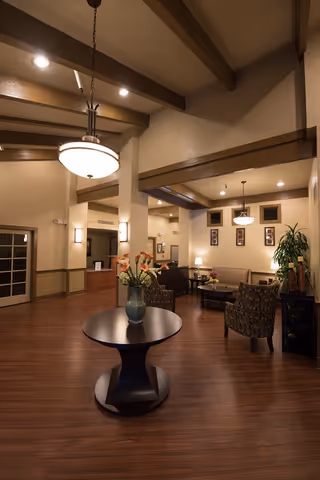 Well-lit lobby lounge with seating areas, a round table with a vase of flowers, pendant lights, and exposed ceiling beams.