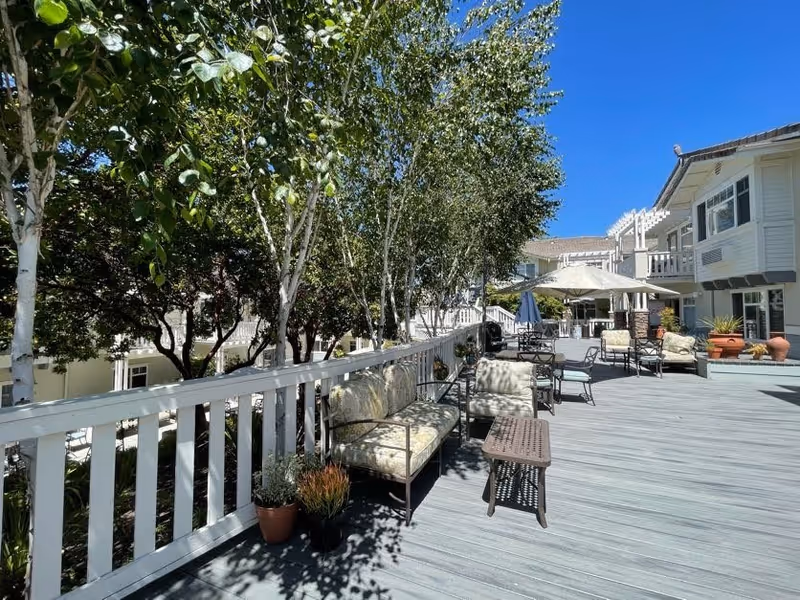 Outdoor deck area at Fremont Hills with cushioned metal chairs and small tables arranged along a white wooden railing. Several potted plants are placed near the railing. Trees provide shade and a clear blue sky is visible above. The building exterior is light-colored with windows and balconies.
