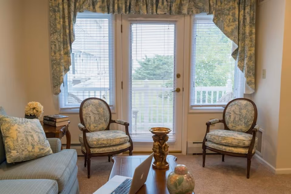 A cozy living room with two upholstered armchairs and a sofa, all featuring a matching blue and white toile pattern. The room has a wooden coffee table with decorative items on it, and a side table with a stack of books and a flower arrangement. Large windows and a glass door with blinds let in natural light and offer a view of a porch and greenery outside.