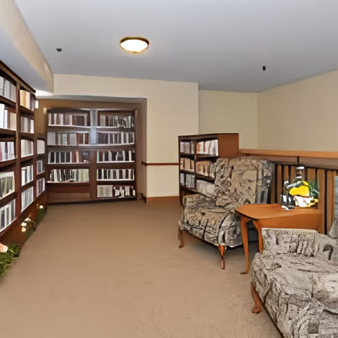 A quiet reading or sitting area in a senior living facility with bookshelves filled with books along the walls, two upholstered armchairs with patterned fabric, and a wooden side table with a lamp on it.