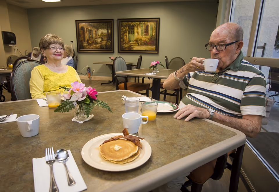 Two elderly people sitting at a dining table in a senior living facility. One woman in a yellow top is smiling, and an elderly man wearing glasses and a striped shirt is drinking from a white cup. On the table are plates with pancakes and bacon, glasses of orange juice, and a small vase with pink flowers. The room has framed paintings on the wall and additional tables and chairs in the background.