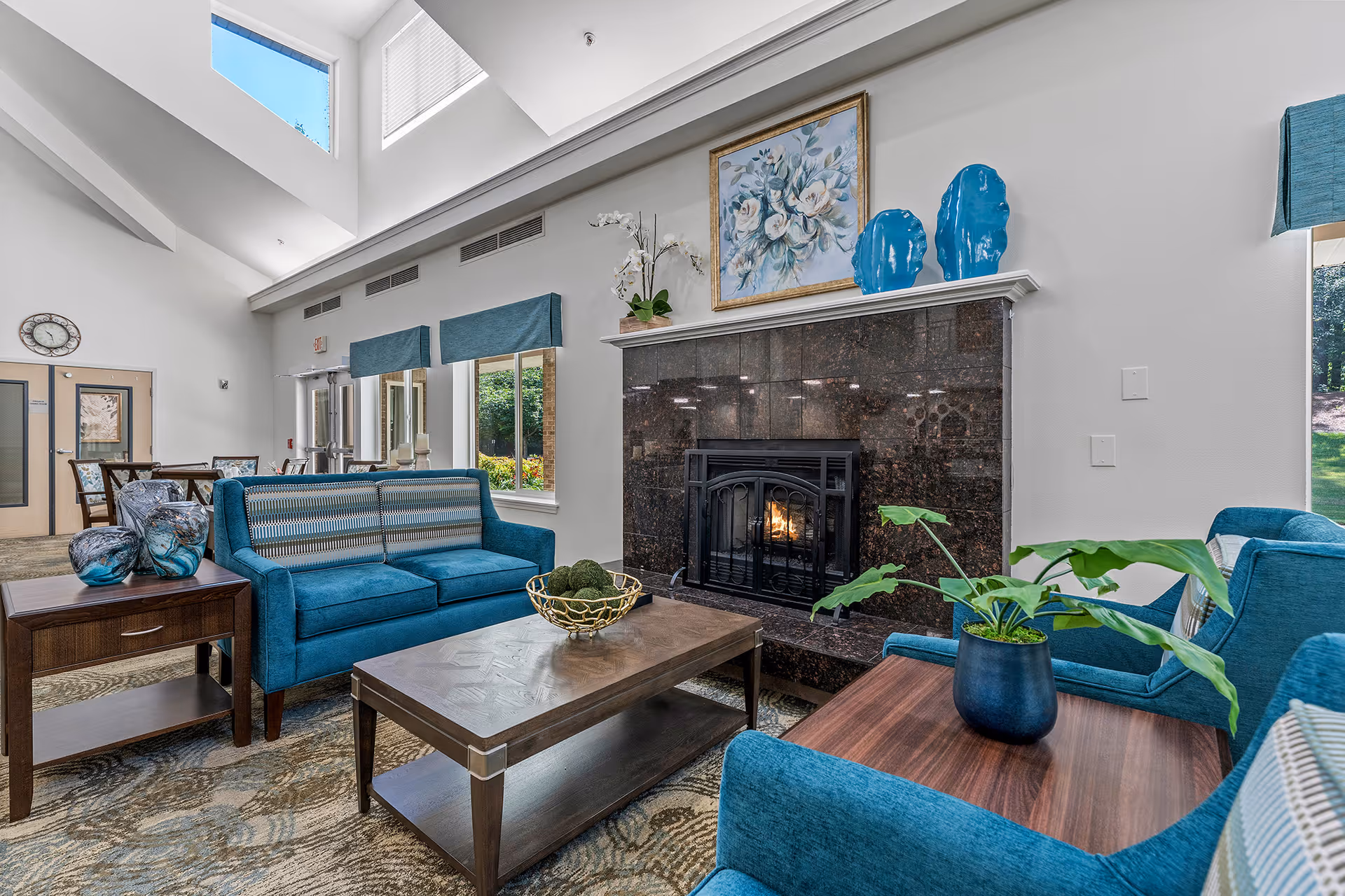 A bright and spacious living room area with high ceilings and large windows letting in natural light. The room features a dark marble fireplace with a fire burning inside, decorated with blue vases and a floral painting above it. There are blue upholstered sofas and armchairs arranged around a wooden coffee table with a decorative bowl. A side table with glass vases and a potted plant on another wooden table add to the cozy ambiance. In the background, a dining area with tables and chairs is visible near the entrance doors.