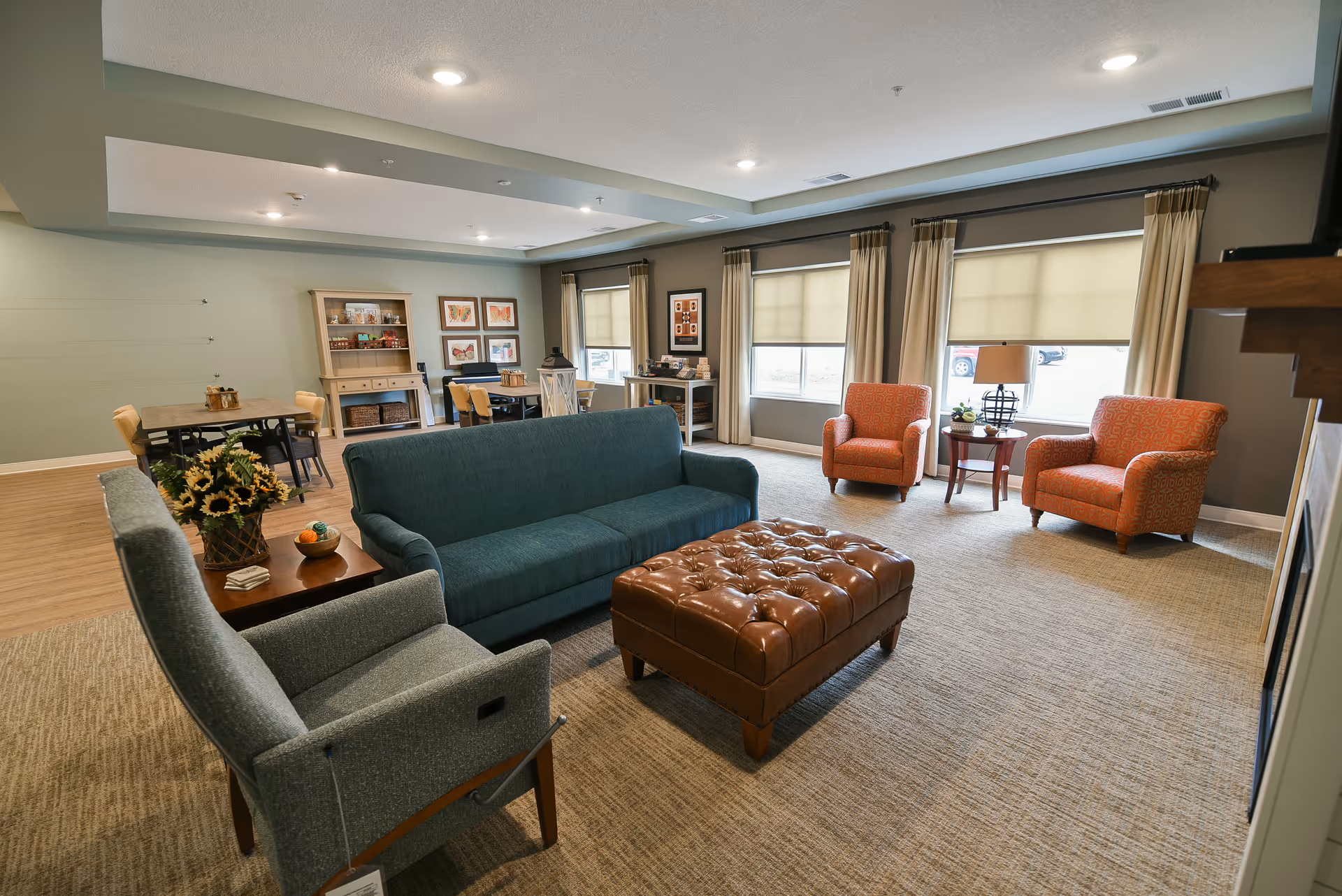 Bright communal living room with sofas, armchairs, a leather ottoman and a dining area in the background.