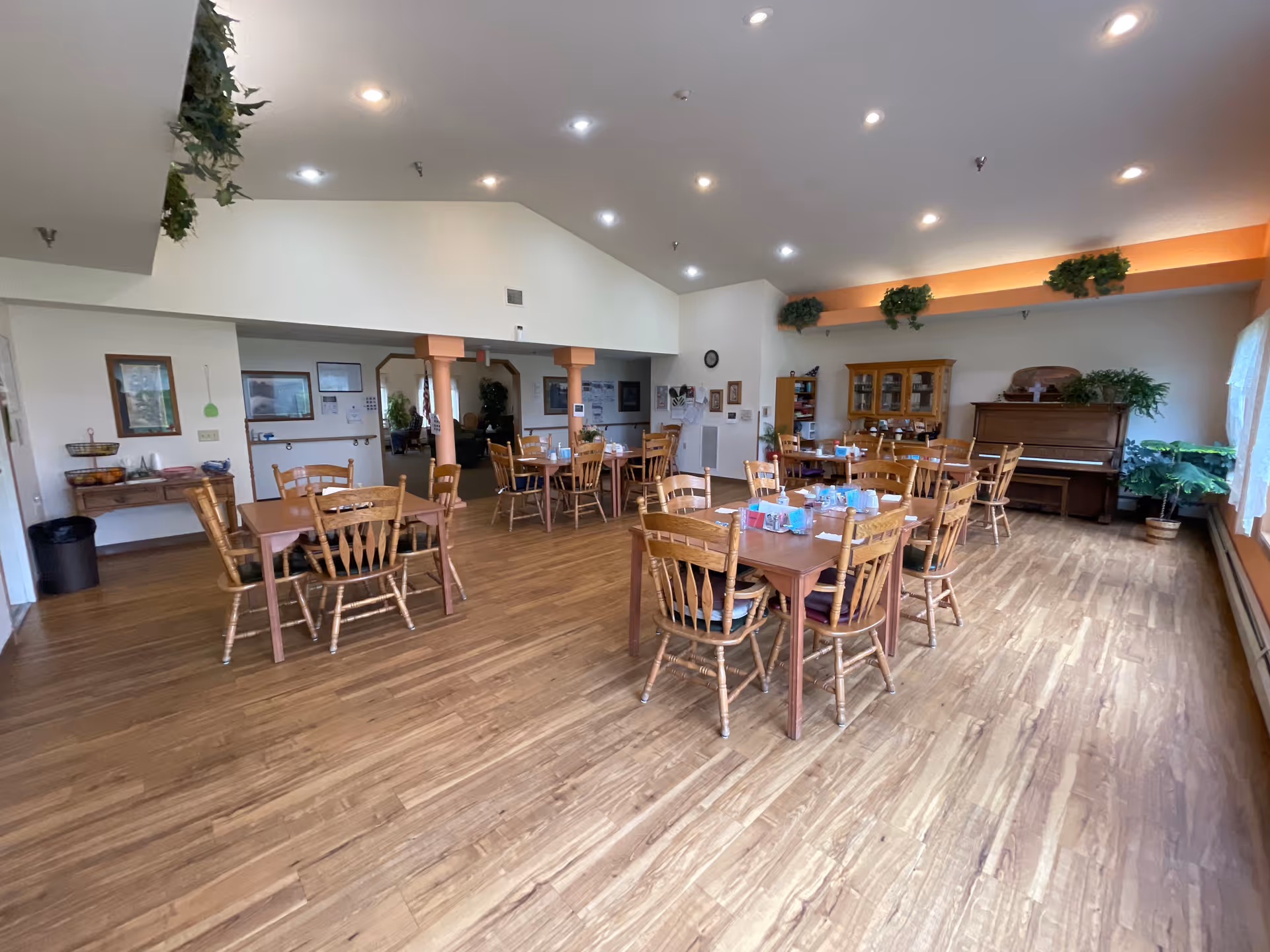 A spacious dining room with wooden floors and multiple wooden tables and chairs arranged neatly. The room has high ceilings with recessed lighting and some green plants placed on shelves and near windows. A piano and a wooden cabinet are visible against one wall, and large windows with sheer curtains allow natural light to fill the space.
