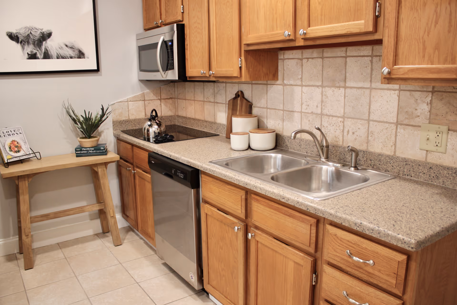 A kitchen area with wooden cabinets, a double stainless steel sink, a dishwasher, an electric stovetop with a kettle, and a microwave mounted above. A small wooden table holds a plant, two books, and a cookbook stand with a cookbook. The backsplash is tiled, and a framed black and white picture of a cow hangs on the wall.