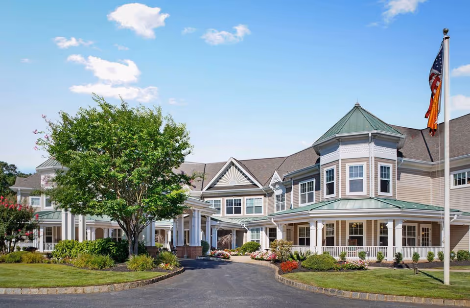 Front exterior of a two-story senior living building with a covered entrance, landscaped grounds, and flags by a circular driveway.