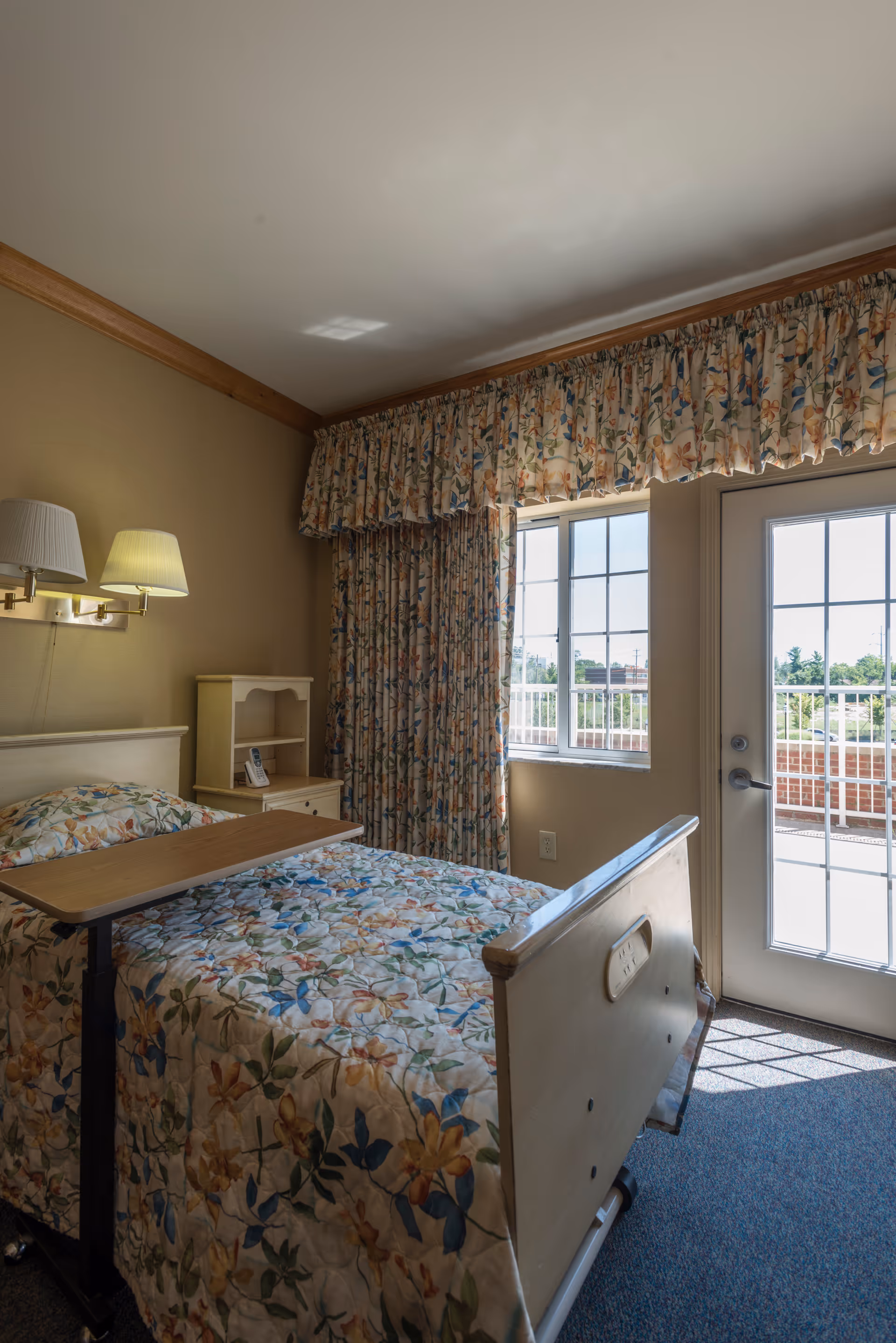 A small, cozy bedroom in a skilled nursing facility with a single bed covered in floral bedding. There is a bedside table with a phone, a wall-mounted lamp above the bed, floral curtains covering a window and a glass door leading outside. The room has beige walls and carpeted floor.