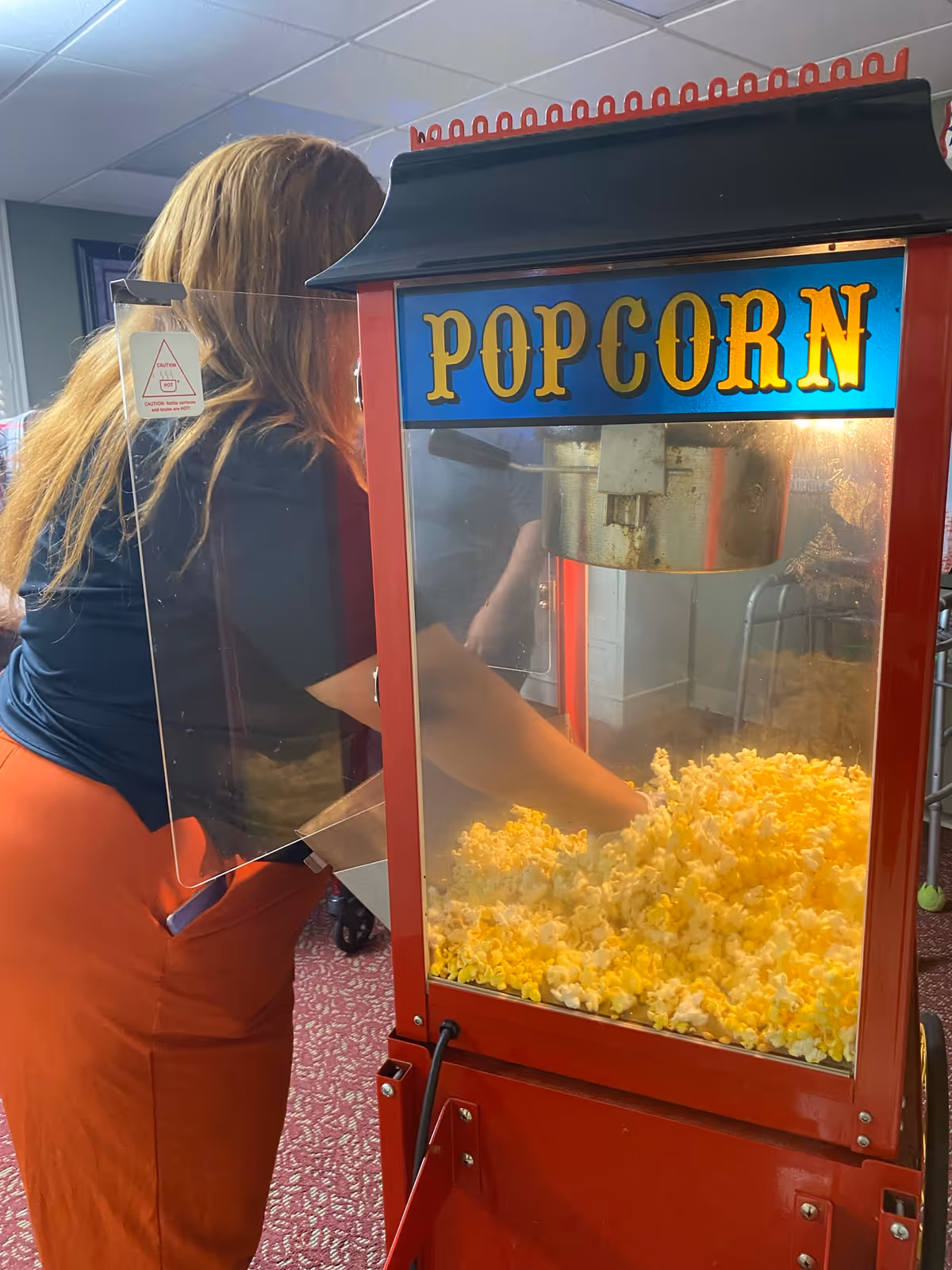 A person with long hair wearing an orange skirt and black top is reaching into a red popcorn machine filled with freshly popped popcorn inside a room with carpeted floor and ceiling tiles.