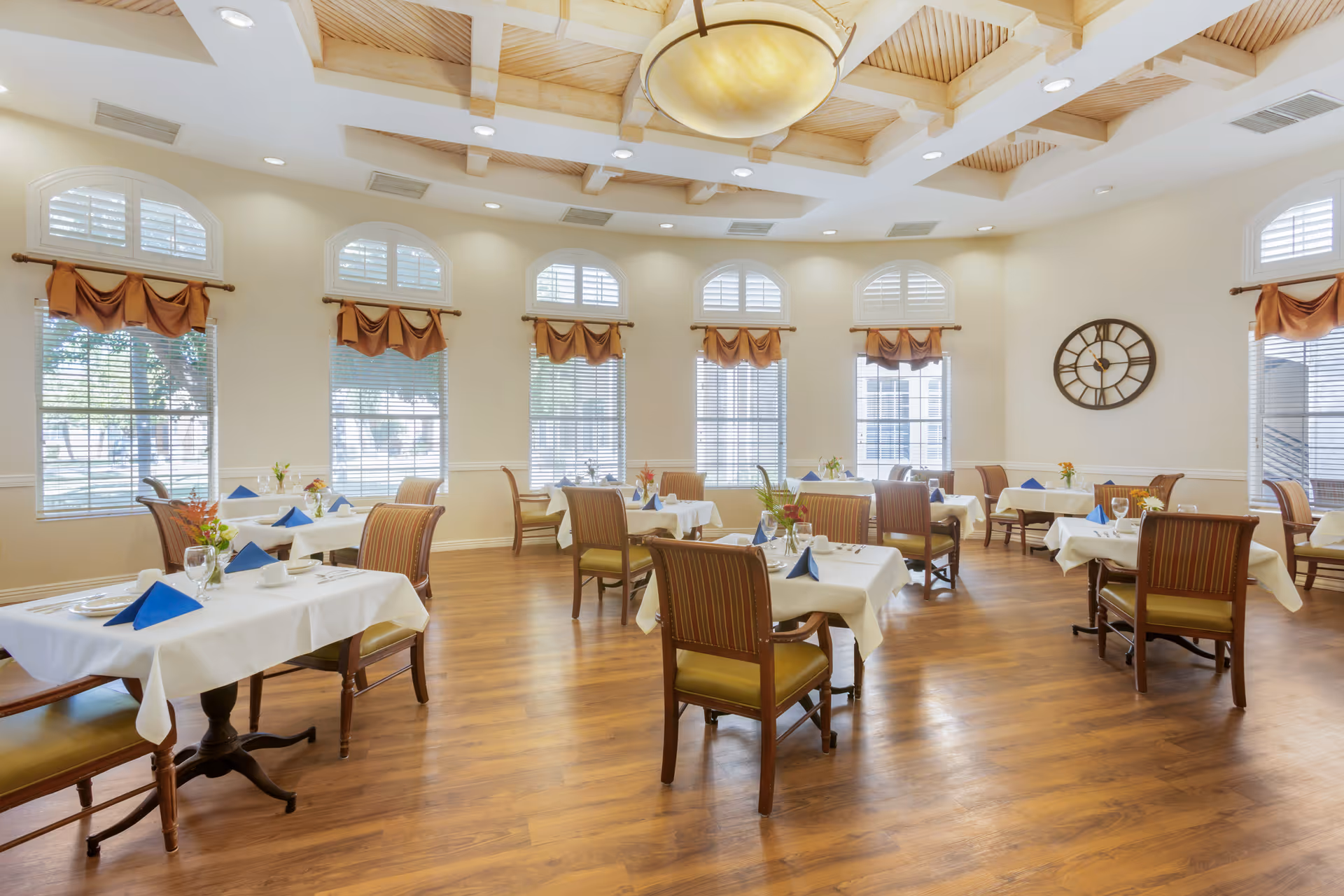 A bright and spacious dining room with wooden floors, multiple tables covered with white tablecloths, each set with blue napkins, glassware, and small floral centerpieces. The room features large windows with white shutters and brown valances, a decorative ceiling with wooden beams, recessed lighting, and a large round wall clock.