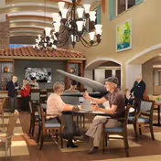 Two elderly people sitting at a table in a spacious, well-lit common area with high ceilings and chandeliers. There is a grand piano in the background, a bar area with shelves and bottles, and other people socializing. The room has warm tones and decorative artwork on the walls.