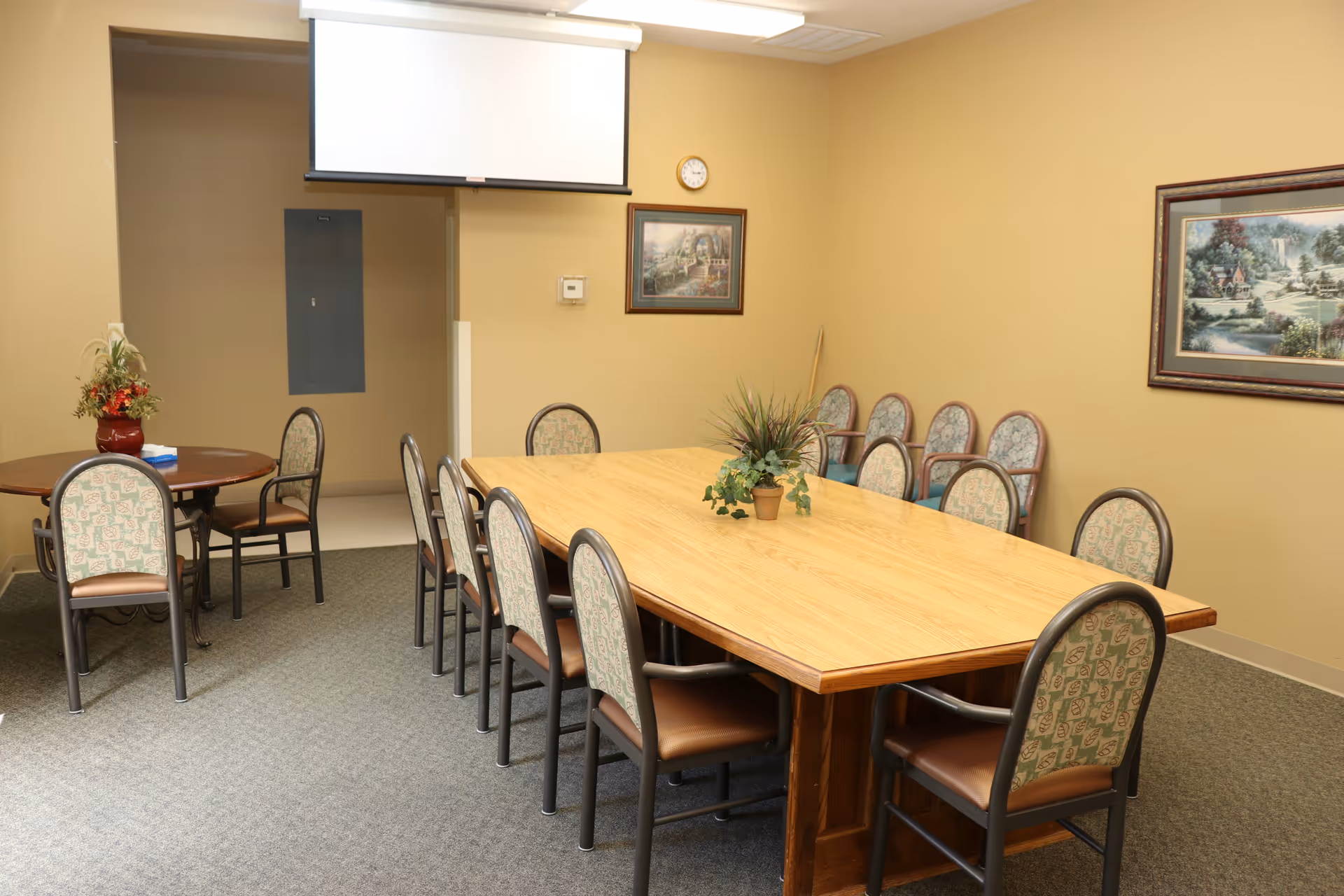A meeting/dining room with a large wooden table surrounded by chairs, a small round table with a flower arrangement, framed paintings, and a ceiling-mounted projection screen.