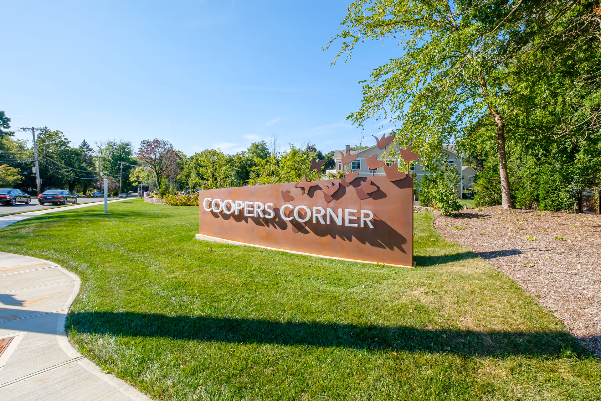 A landscaped entrance with a metal sign reading "COOPERS CORNER" on a grassy lawn with trees, sidewalk, and a building in the background.