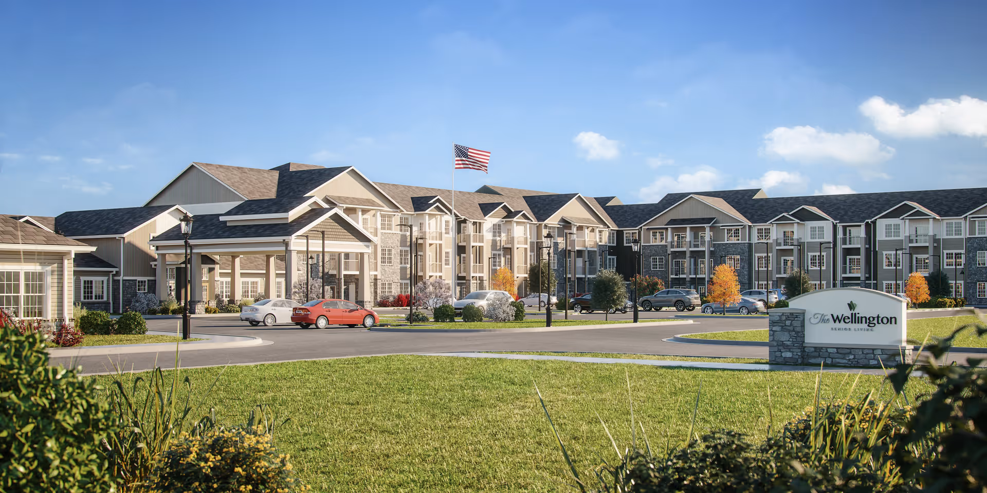 Exterior view of The Wellington Senior Living facility showing a large multi-story building with multiple windows and balconies, a driveway with parked cars, an American flag on a flagpole, and a landscaped lawn with bushes and trees under a blue sky with some clouds.