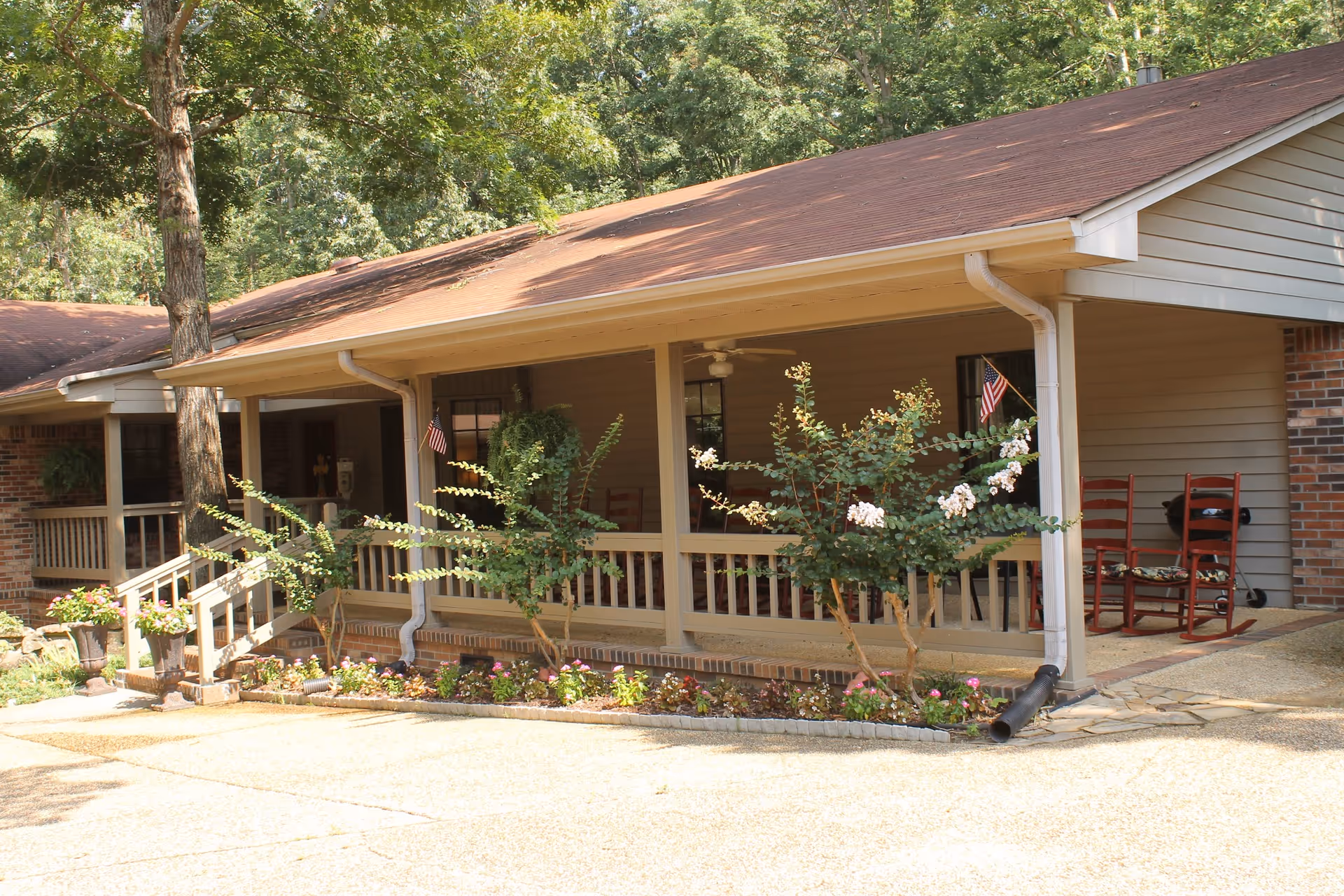 Front porch of a single-story building with a brown roof and beige siding, featuring a small garden with flowering plants and shrubs in front. The porch has a railing, two American flags, and red rocking chairs. Trees and greenery surround the area.