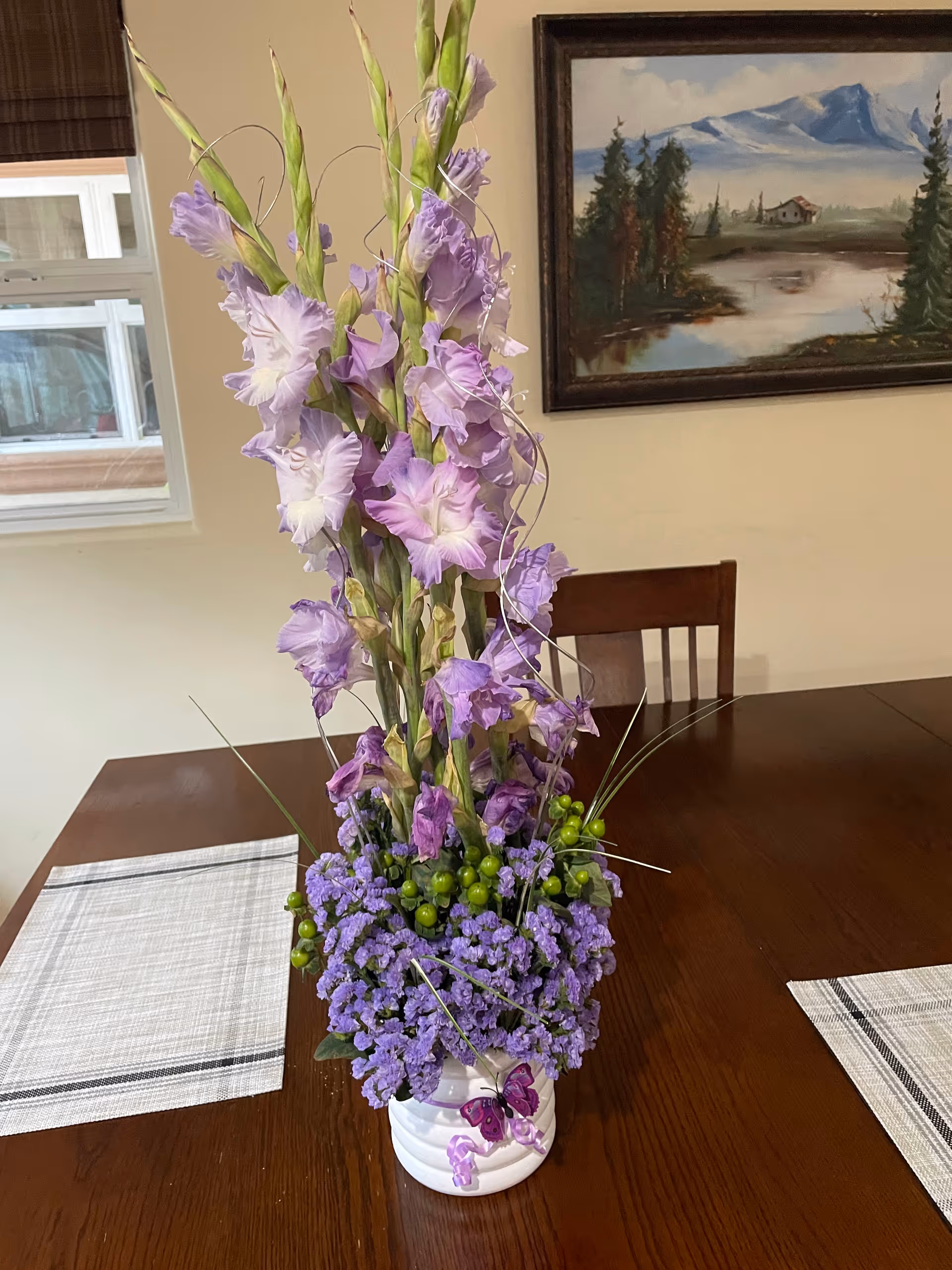 A floral arrangement with tall light purple gladiolus flowers, small purple flowers, and green berries in a white vase with a purple butterfly decoration, placed on a wooden dining table with two white and gray placemats. In the background, there is a window with brown blinds and a framed landscape painting of mountains, trees, and a lake on a beige wall.