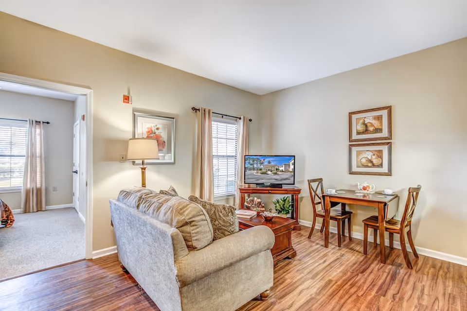 Bright living room with a sofa facing a TV, a small dining table with chairs, and a doorway leading to a bedroom.