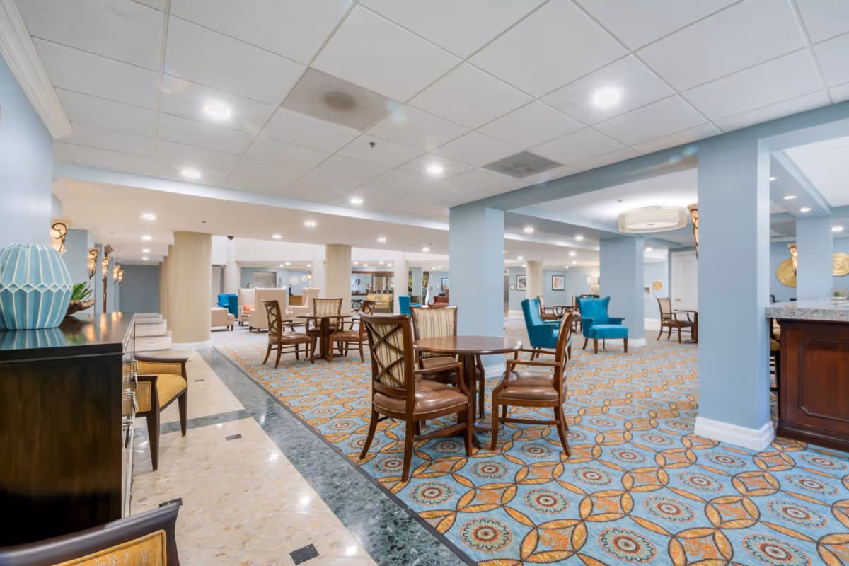 Bright communal dining and lounge area with patterned carpet, round wooden tables, and teal accent chairs.