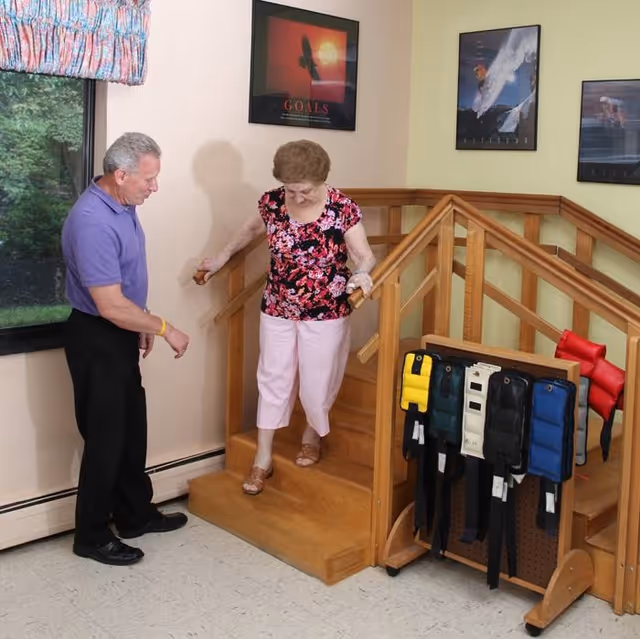 An elderly woman is walking down a small wooden staircase while holding onto the handrails on both sides. A man stands nearby, watching and ready to assist. The room has light-colored walls with three framed motivational posters, a window with a floral curtain, and a rack holding various weighted belts or straps.