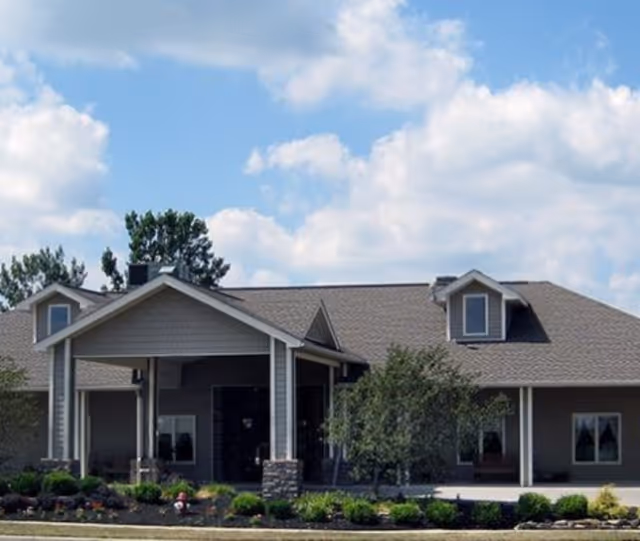 Exterior view of a single-story assisted living facility building with a covered entrance, surrounded by landscaping and trees under a partly cloudy sky.