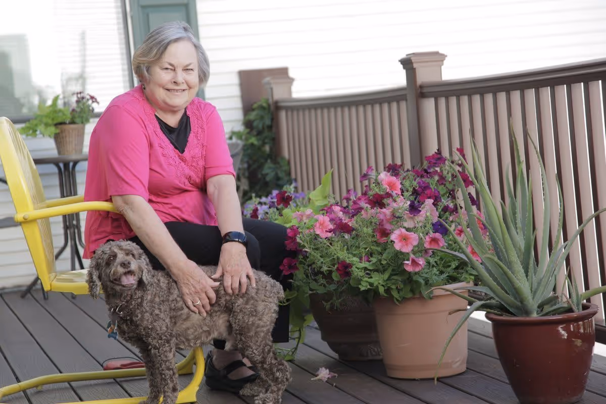 An elderly woman wearing a pink shirt sitting on a yellow chair on a wooden deck, smiling and petting a small curly-haired dog. There are potted plants with colorful flowers and an aloe plant next to her, with a railing and part of a building visible in the background.