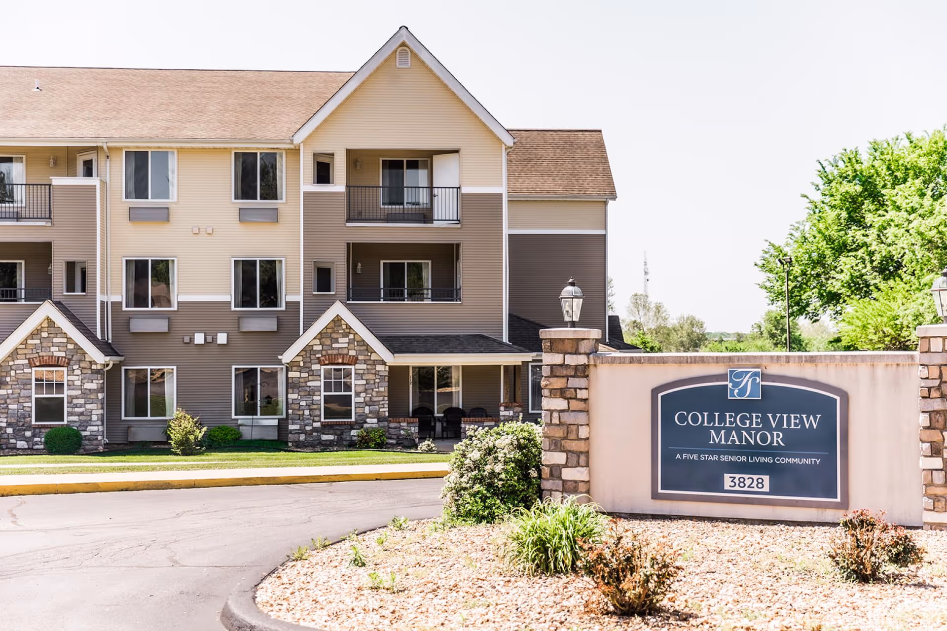 Exterior view of College View Manor, a senior living community building with beige and brown siding, stone accents, balconies, and a landscaped entrance with a sign displaying the facility name and address 3828.