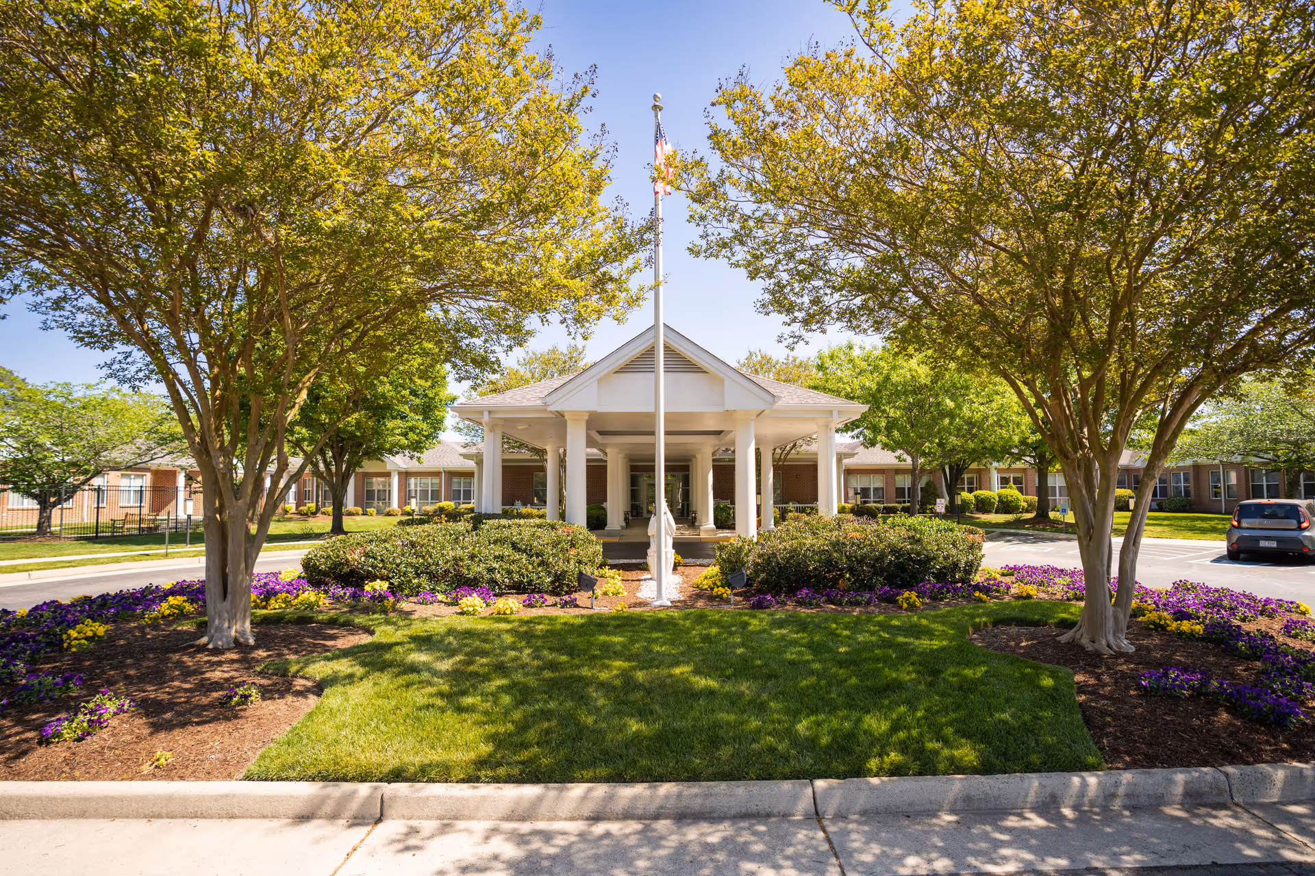 Front entrance of a single-story senior living building with a covered portico, flagpole, and landscaped lawn and trees.