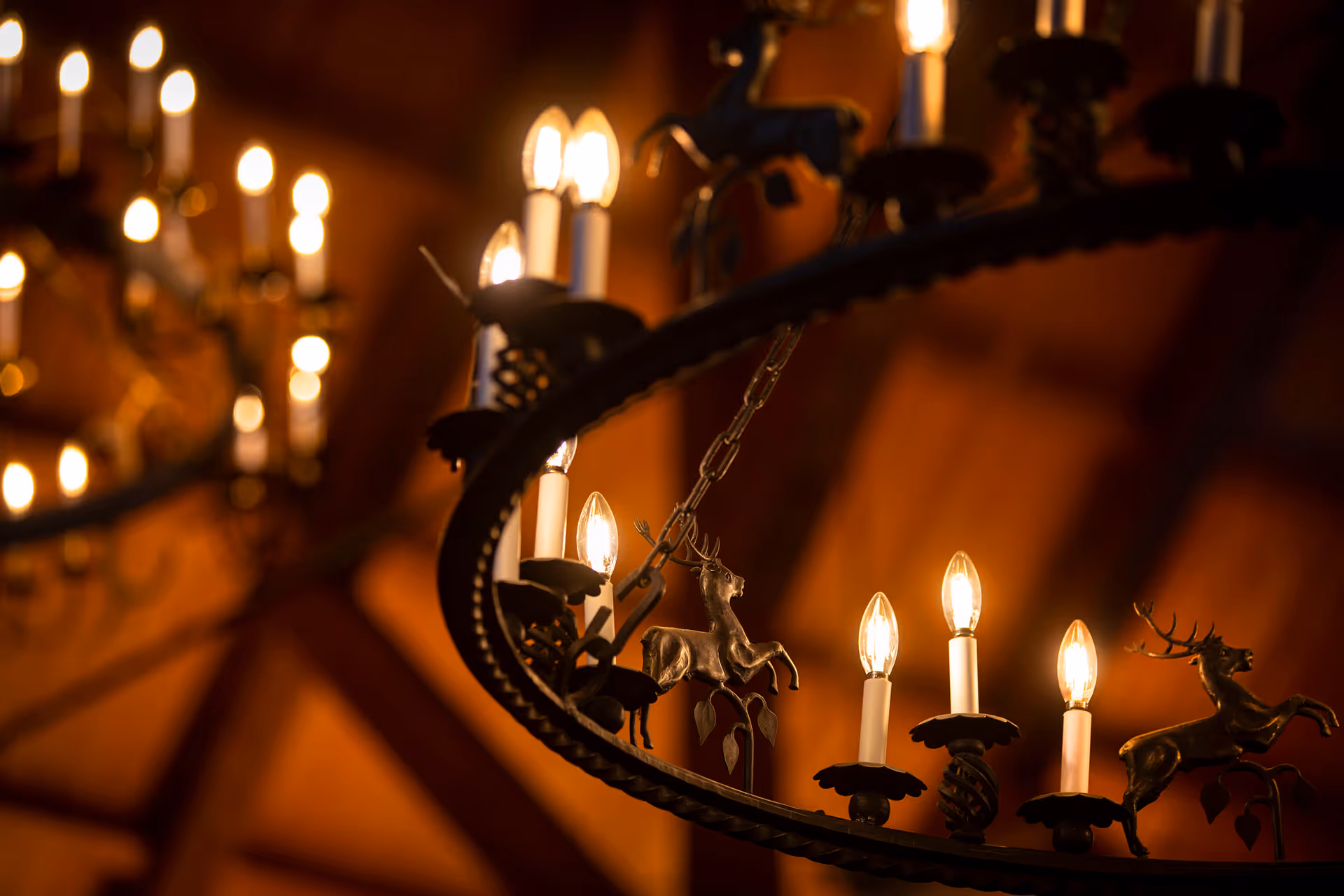 Ornate chandelier with candle-shaped bulbs and small deer figurines hanging beneath a warm, wooden-beamed ceiling.