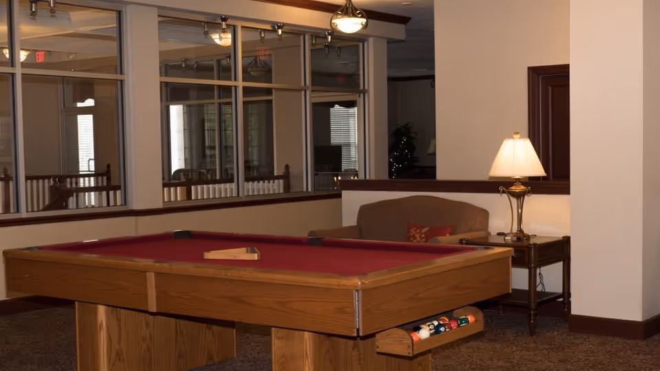 Interior view of a common area featuring a wooden pool table with a red felt surface and pool balls stored in a rack underneath. Behind the pool table is a brown upholstered sofa with a decorative pillow, next to a wooden side table with a lit table lamp. Large windows with multiple panes reflect the room's lighting and furnishings.