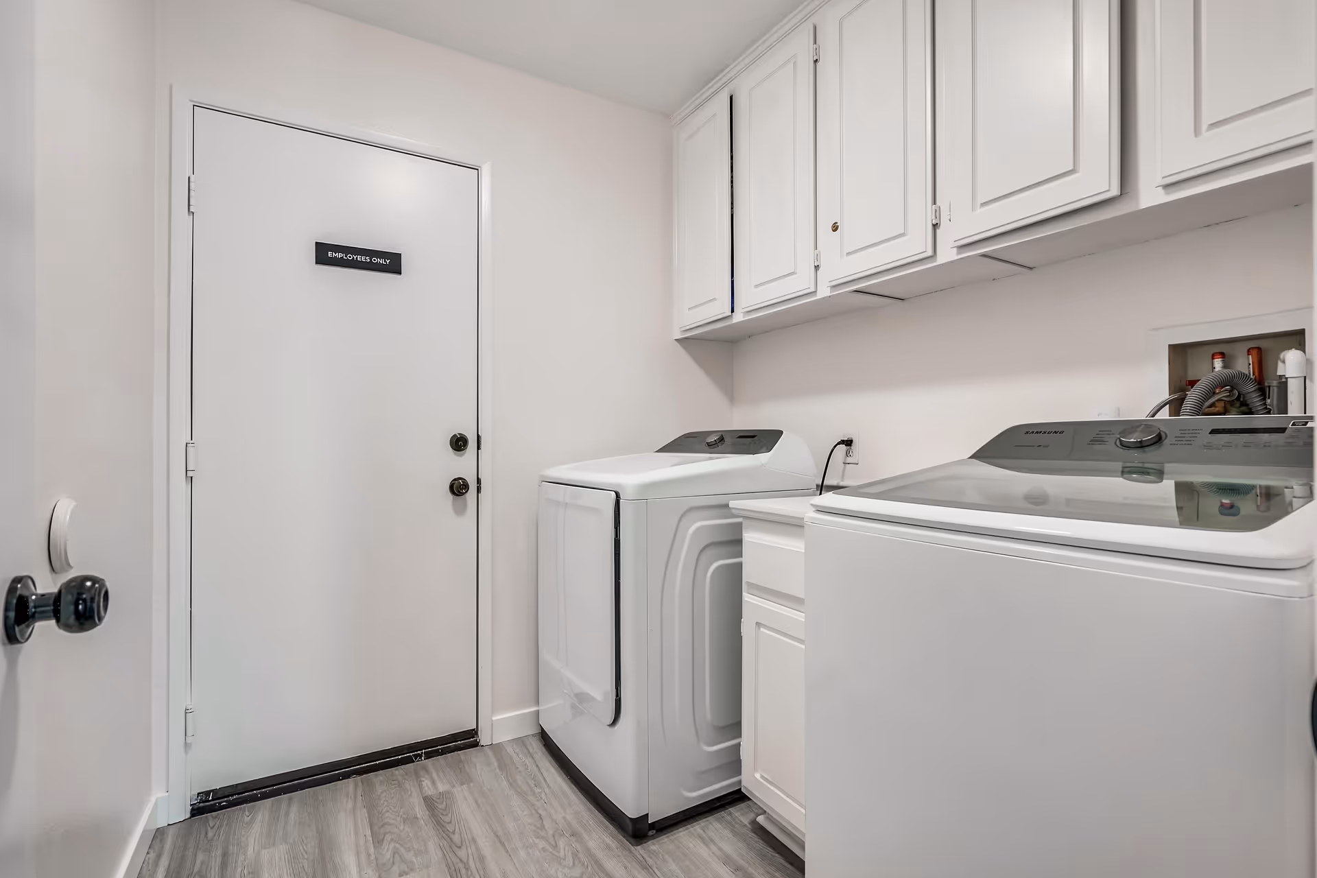 Bright laundry room with a washer and dryer, white cabinets overhead, and a door labeled "EMPLOYEES ONLY".