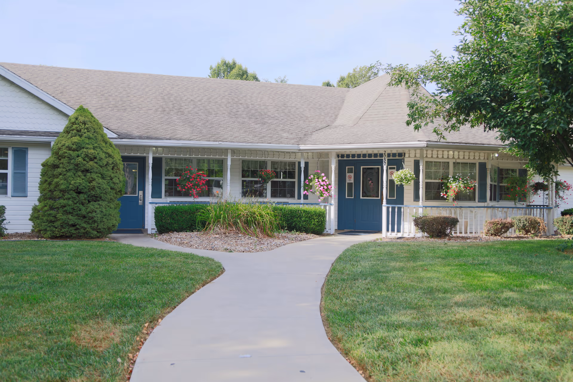 Front exterior of a single-story assisted living building with a curved walkway, blue doors, porch and hanging flower baskets.