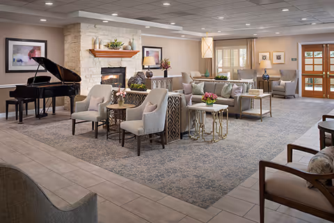 Spacious senior living facility common area with comfortable seating including armchairs and sofas arranged around a stone fireplace. A grand piano is positioned on the left side of the room. The room features soft lighting, framed artwork on the walls, and large windows with curtains allowing natural light. The floor is a combination of tiled and carpeted areas, and there are small tables with flower arrangements throughout the space.
