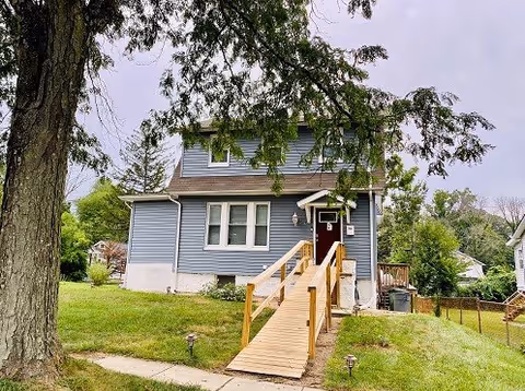 Front view of a two-story blue house with a wooden accessibility ramp, grassy yard, and a large tree in front.