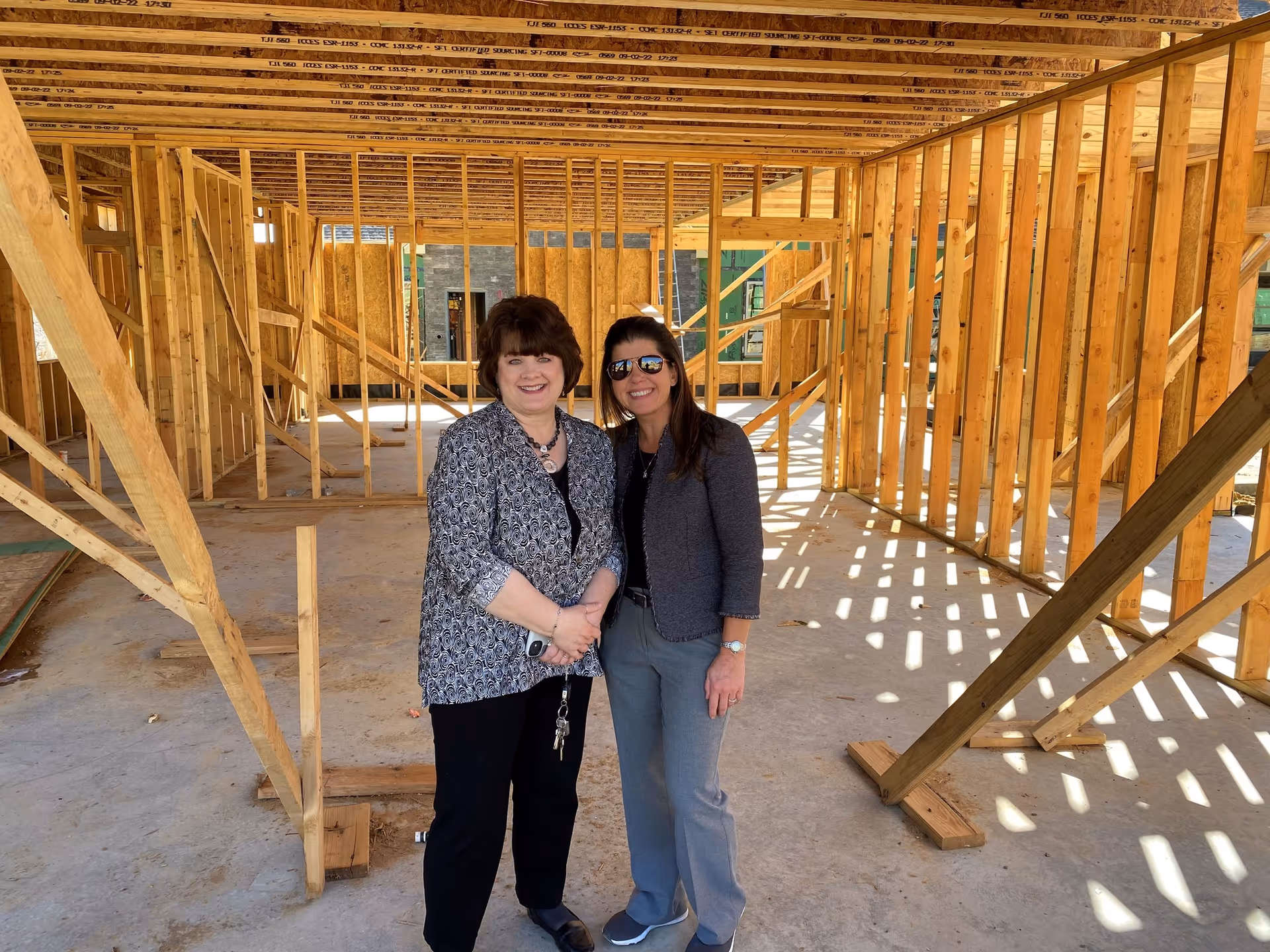 Two women standing inside a wooden frame structure of a building under construction, smiling at the camera. The framing shows exposed wooden beams and studs with sunlight casting shadows on the concrete floor.