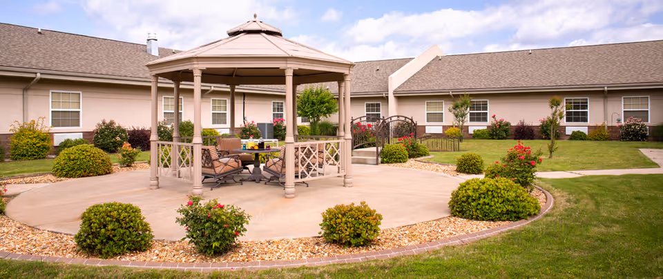 Outdoor courtyard with a central gazebo, seating, and landscaped shrubs in front of a single-story nursing facility building.
