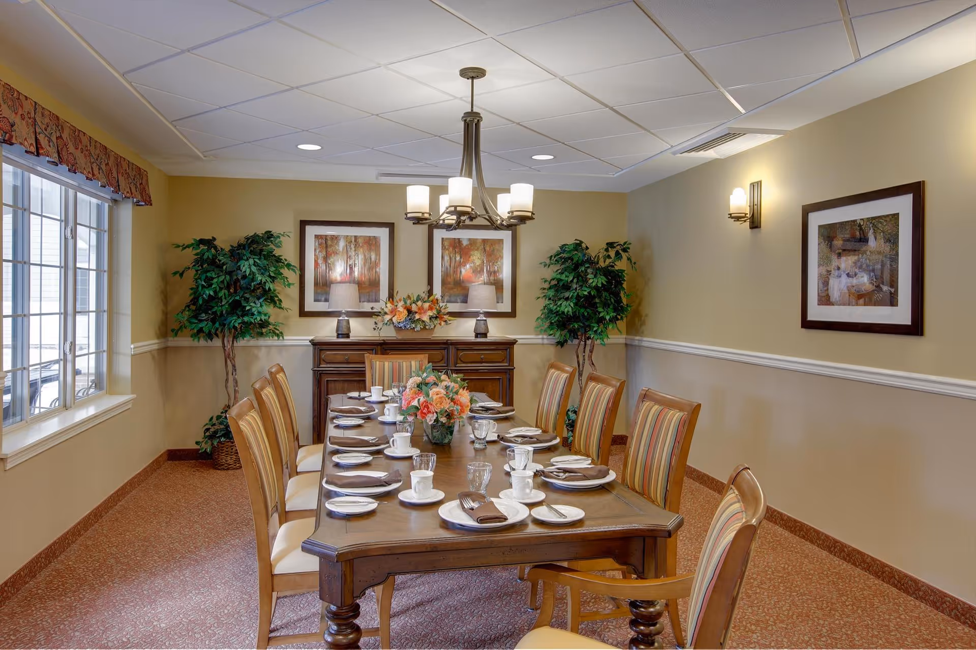 A formal dining room with a long wooden table set for ten people with plates, cups, glasses, and napkins. The room has beige walls with white trim, two framed paintings, two potted plants, a sideboard with lamps and a flower arrangement, and a window with a valance on the left side.