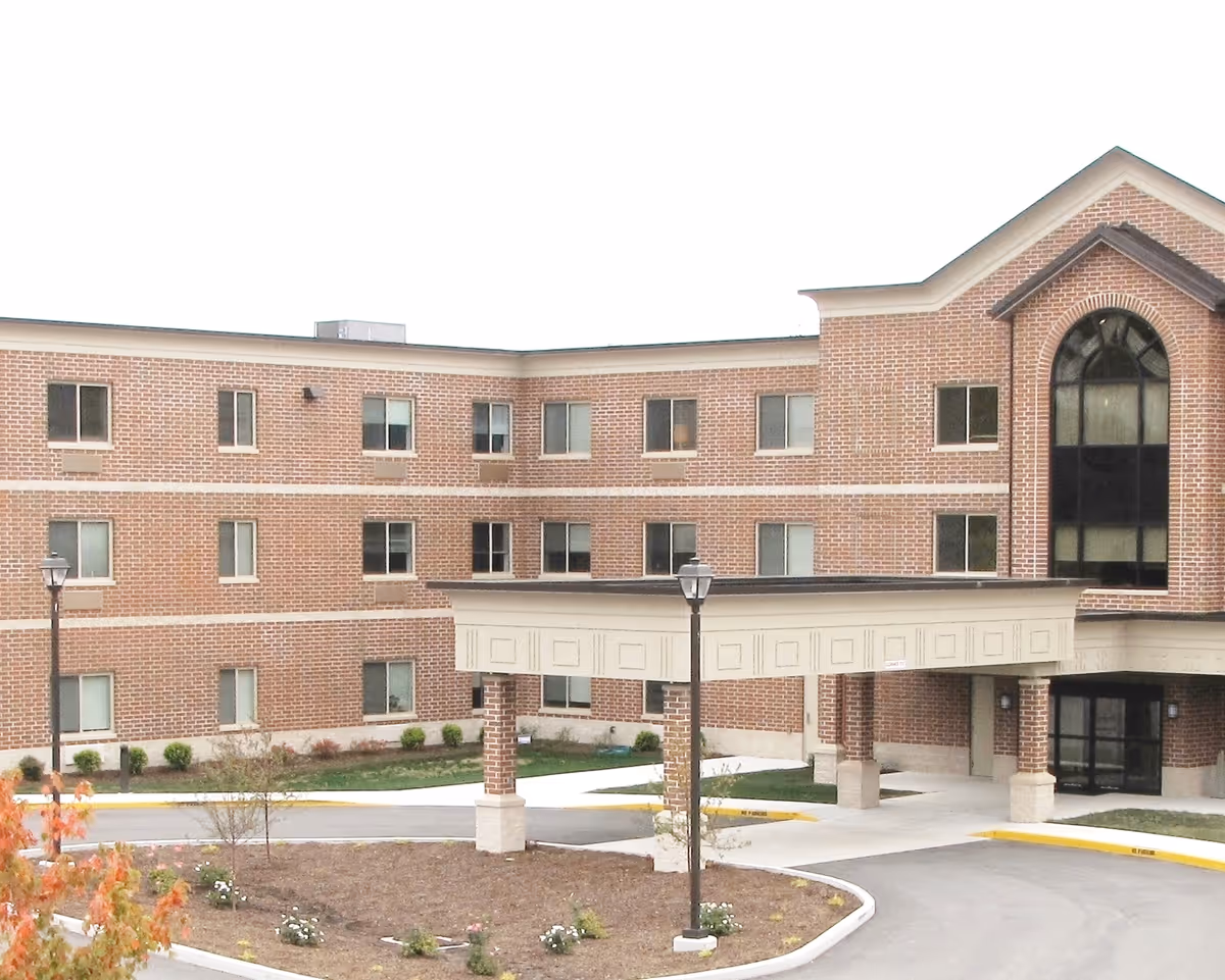 Exterior view of a three-story brick building with multiple windows and a covered entrance supported by brick columns. There are small landscaped areas with plants and a curved driveway in front of the entrance.
