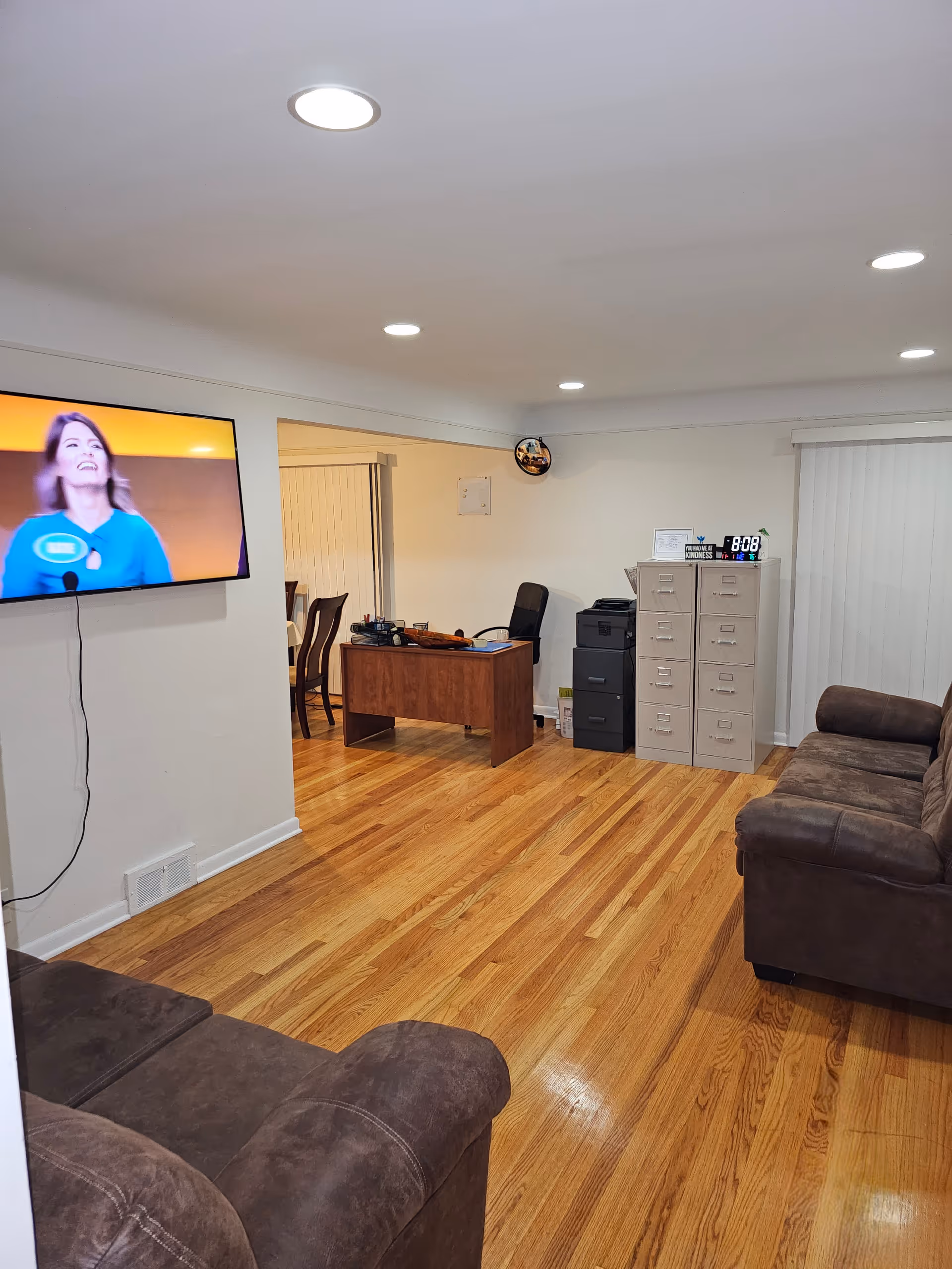 A room with wooden flooring featuring two brown sofas facing each other. A flat-screen TV is mounted on the left wall showing a woman in a blue top. In the background, there is a wooden desk with office supplies, a black office chair, filing cabinets, and a printer. The room is well-lit with recessed ceiling lights.