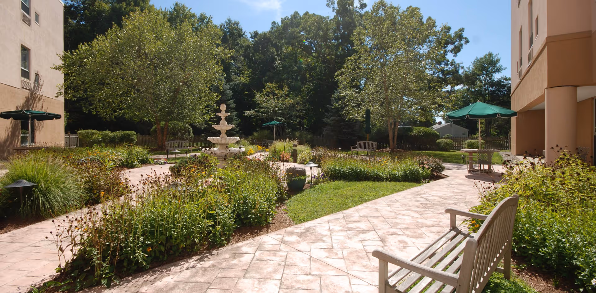 Sunny landscaped courtyard with paved walkways, benches, a central fountain, umbrellas, and surrounding building façades.