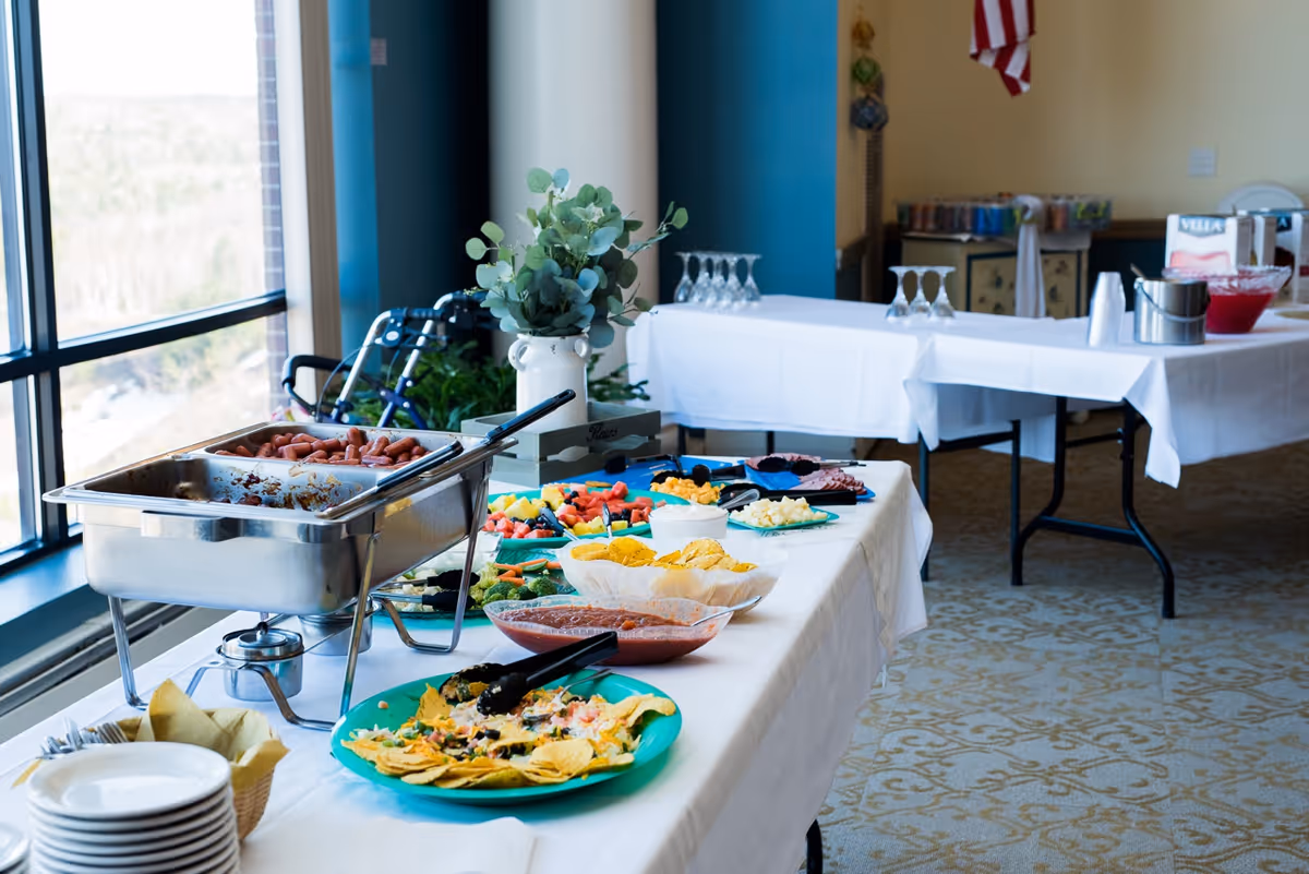 Buffet-style spread of food including nachos, chips and dip, fruit, and hot dogs arranged on tables in a bright dining area with large windows.