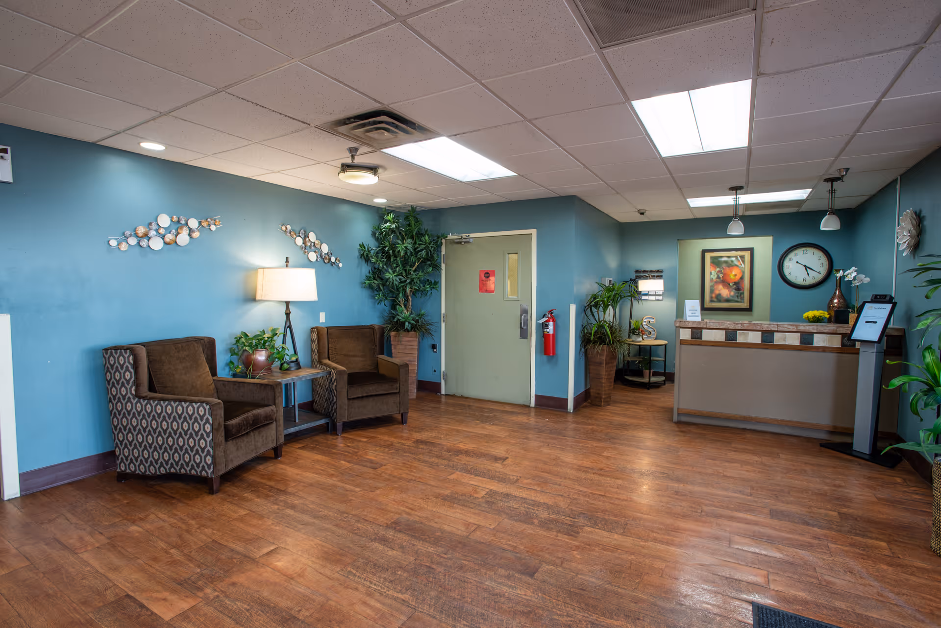 Reception area of Sandstone of Tucson facility with two brown patterned armchairs and a small table with a lamp and plant on the left side, a green door in the center, and a reception desk on the right with a clock, framed artwork, and plants around the room. The walls are painted blue and the floor has wood-like flooring.