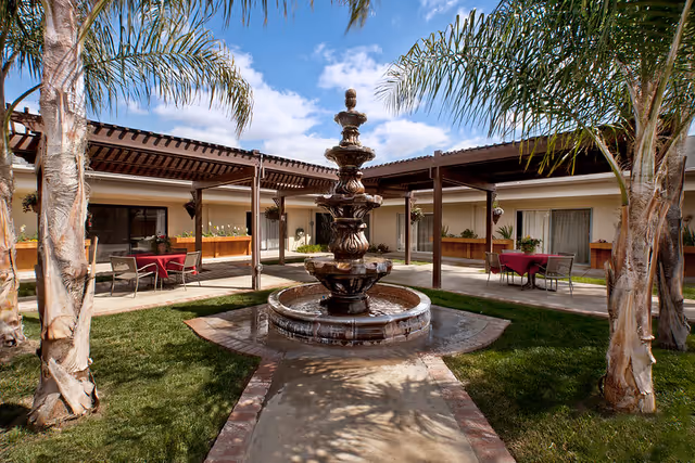 Outdoor courtyard area with a multi-tiered water fountain in the center, surrounded by palm trees and green grass. There are covered walkways with wooden pergolas and tables with chairs and red tablecloths near the building entrances under the pergolas. The sky is blue with some clouds.