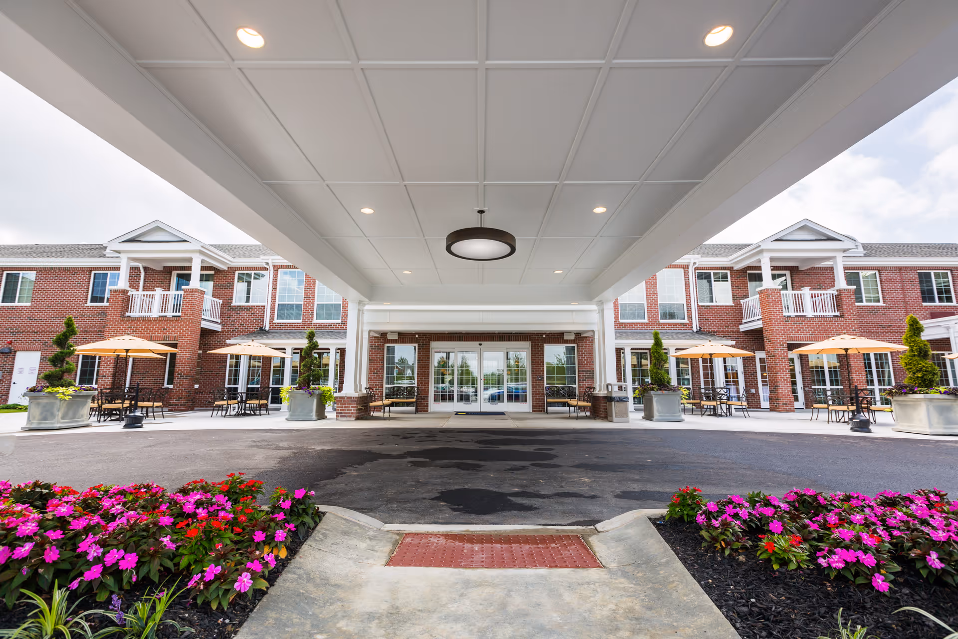 Entrance of a senior living facility with a covered drop-off area, brick building facade, outdoor seating with tables and umbrellas, and flower beds with pink and red flowers in the foreground.