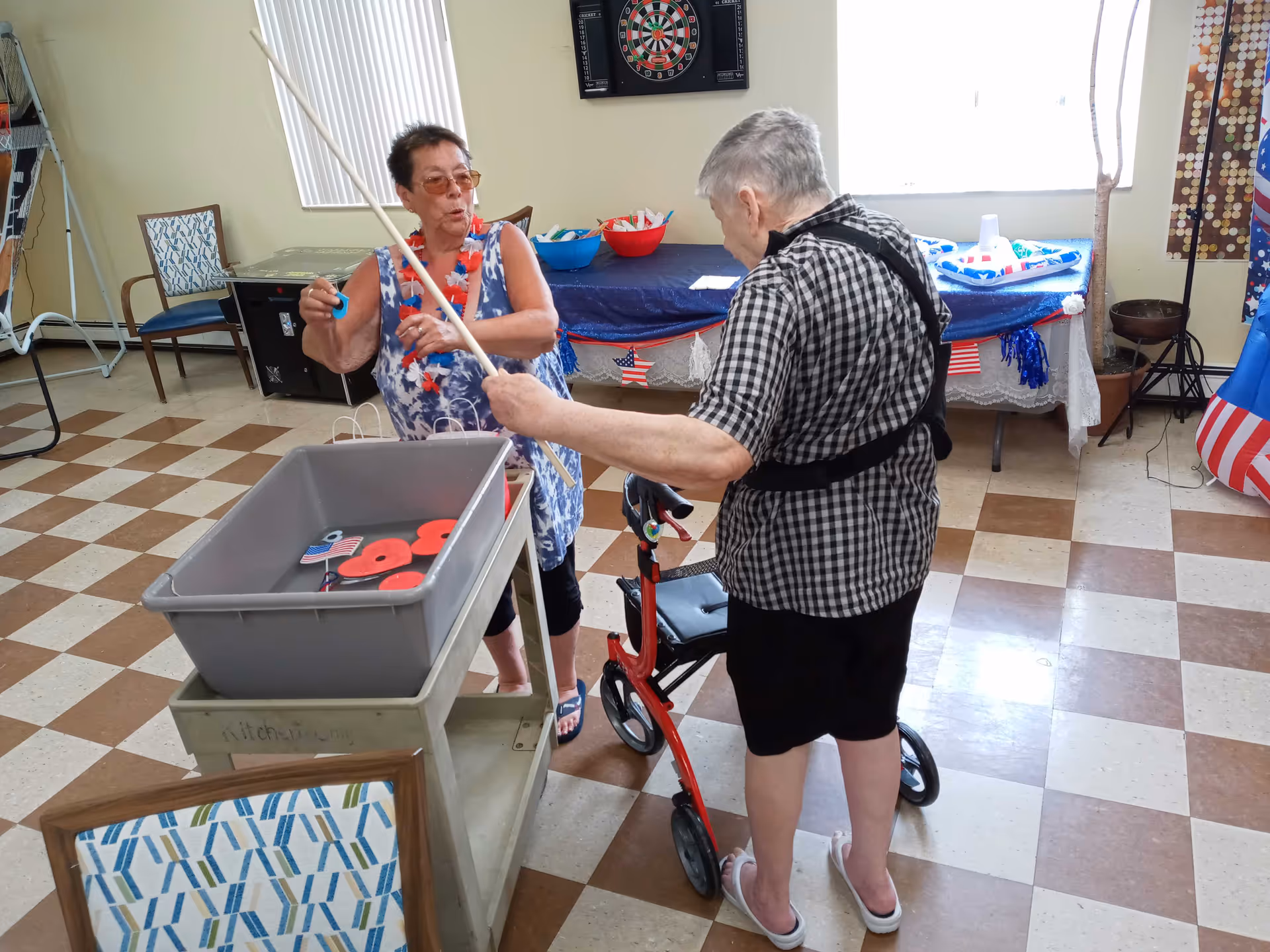 Two elderly women playing a ring toss game indoors. One woman is holding a fishing pole with a ring, while the other woman, using a walker, is holding a stick. The room has a checkered floor, a dartboard on the wall, and a table decorated with patriotic items in the background.