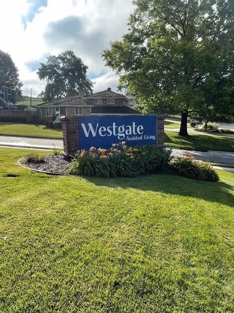A large blue sign with white text reading 'Westgate Assisted Living' is placed on a landscaped grassy area with flowers in front. Behind the sign, there are trees, a road, and a residential-style building under a partly cloudy sky.