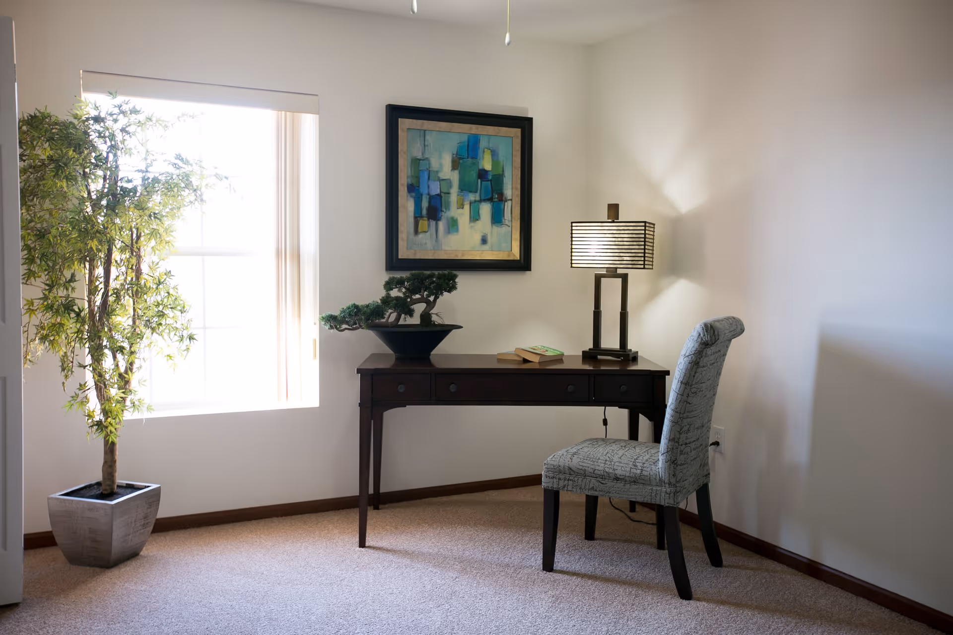 A small corner of a room with a window letting in natural light, a potted plant on the floor, a dark wooden desk with a bonsai tree, two books, and a modern table lamp. A patterned upholstered chair is placed in front of the desk, and an abstract painting hangs on the wall above the desk.