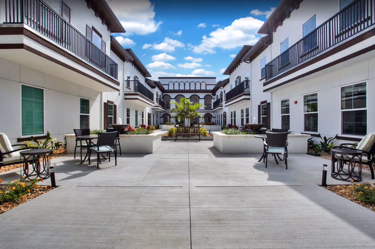 Outdoor courtyard area of a senior living facility with white buildings on both sides featuring balconies. The courtyard has seating areas with chairs and small tables, planters with greenery and flowers, and a bench with a palm tree in the center. The sky is blue with scattered clouds.