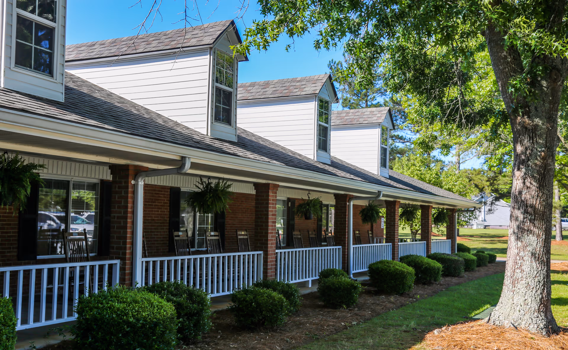 Exterior view of a senior living facility with a covered porch featuring rocking chairs and hanging plants. The building has brick walls, white railings, and dormer windows on the roof. There are green bushes and a large tree in the foreground under a clear blue sky.