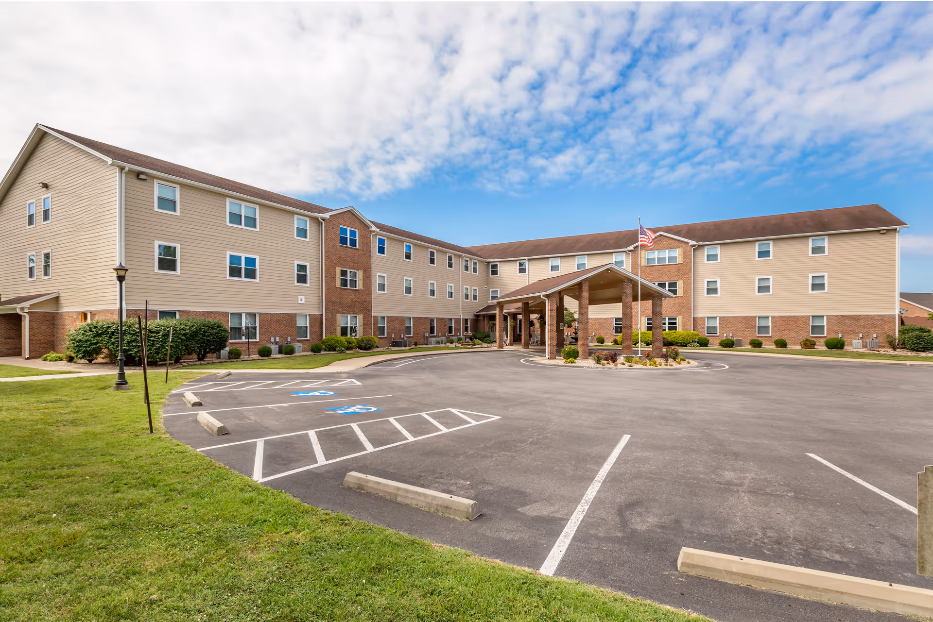 Exterior view of a three-story senior living facility building with beige siding and brick accents. The building has multiple windows and a covered entrance with an American flag on a flagpole. The foreground shows a parking lot with marked handicap parking spaces and green grass on the sides under a partly cloudy blue sky.