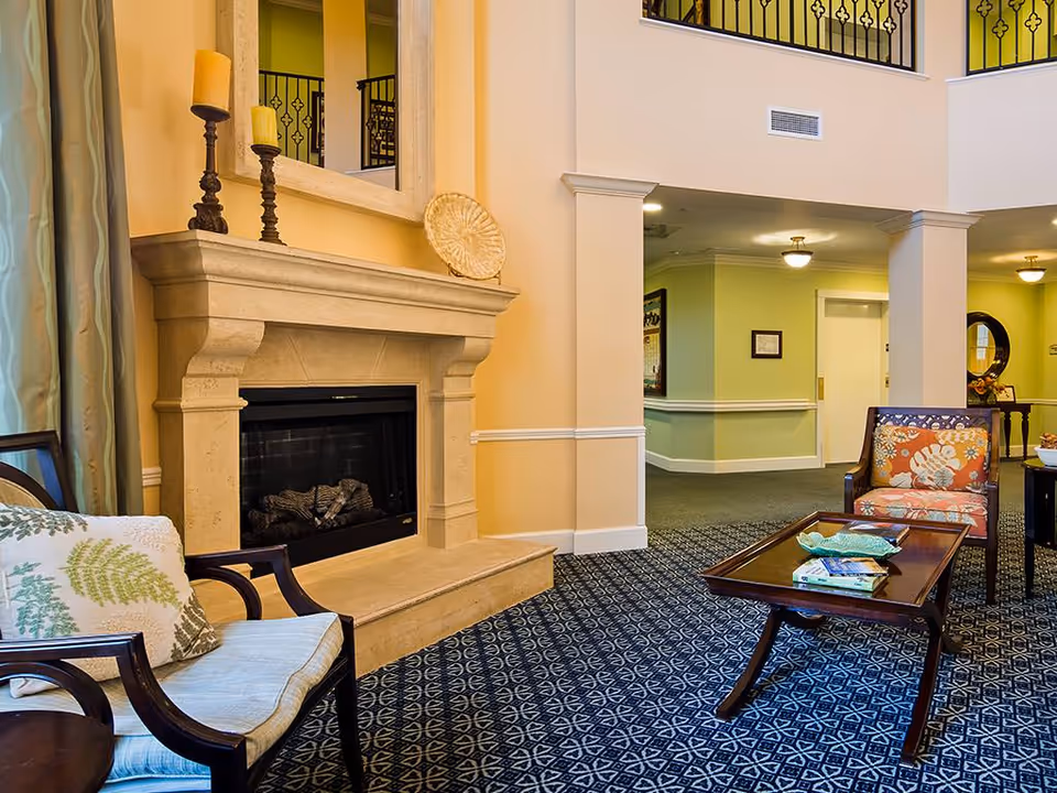 A cozy living room area with a stone fireplace, two armchairs with patterned cushions, a wooden coffee table with decorative items, and a carpeted floor. The walls are painted in warm tones with white trim, and there is a large mirror above the fireplace. The background shows a hallway with green walls and ceiling lights.